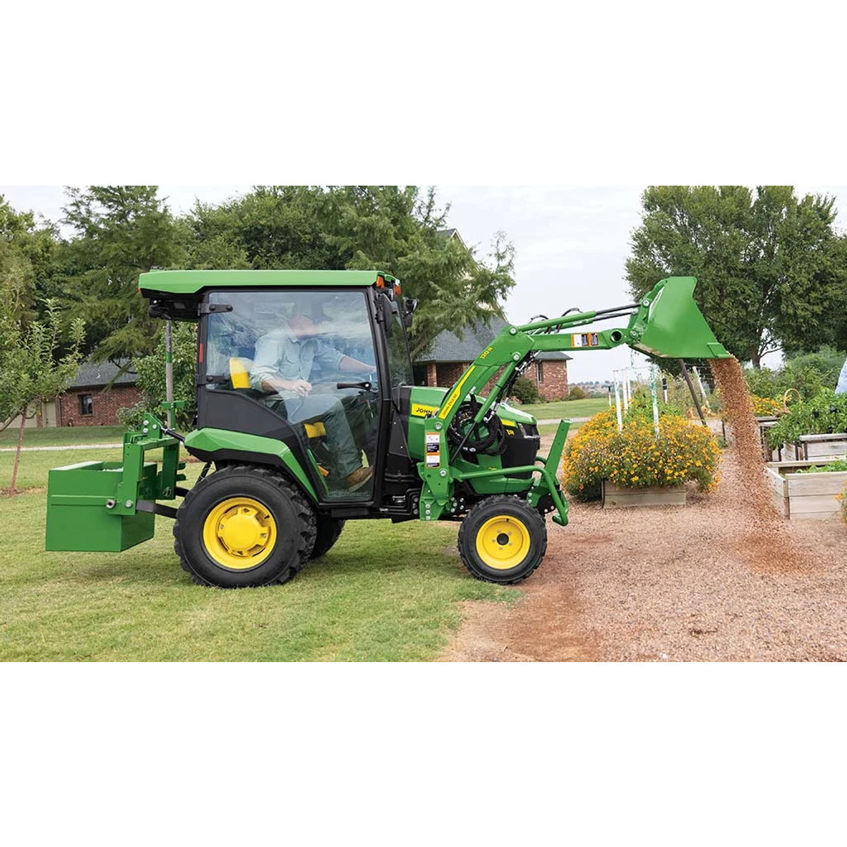 A green compact tractor with a rear ballast box and front loader dumps gravel around some planter boxes.