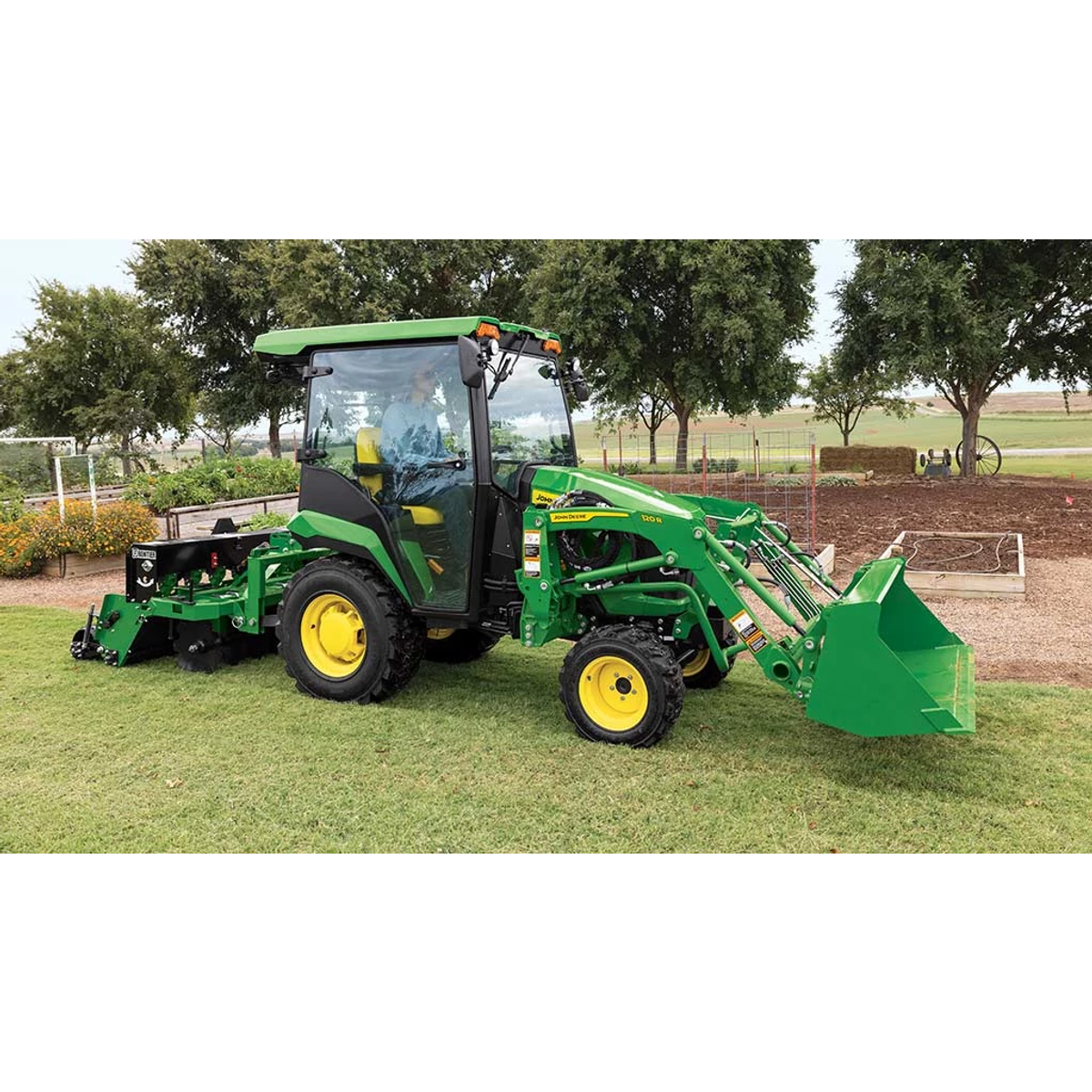 A green compact tractor with a loader and ground seeder drives next to a garden plot.