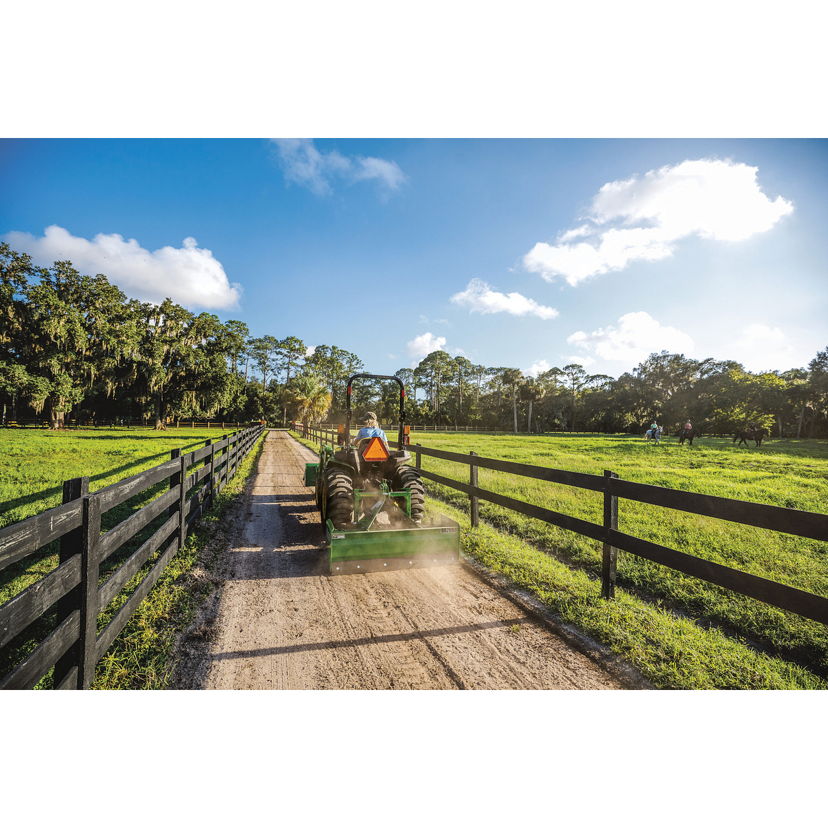 A green tractor levels a dirt lane between wooden fences with a rear land plane, with open grassy fields and trees on both sides under a bright blue sky with scattered clouds