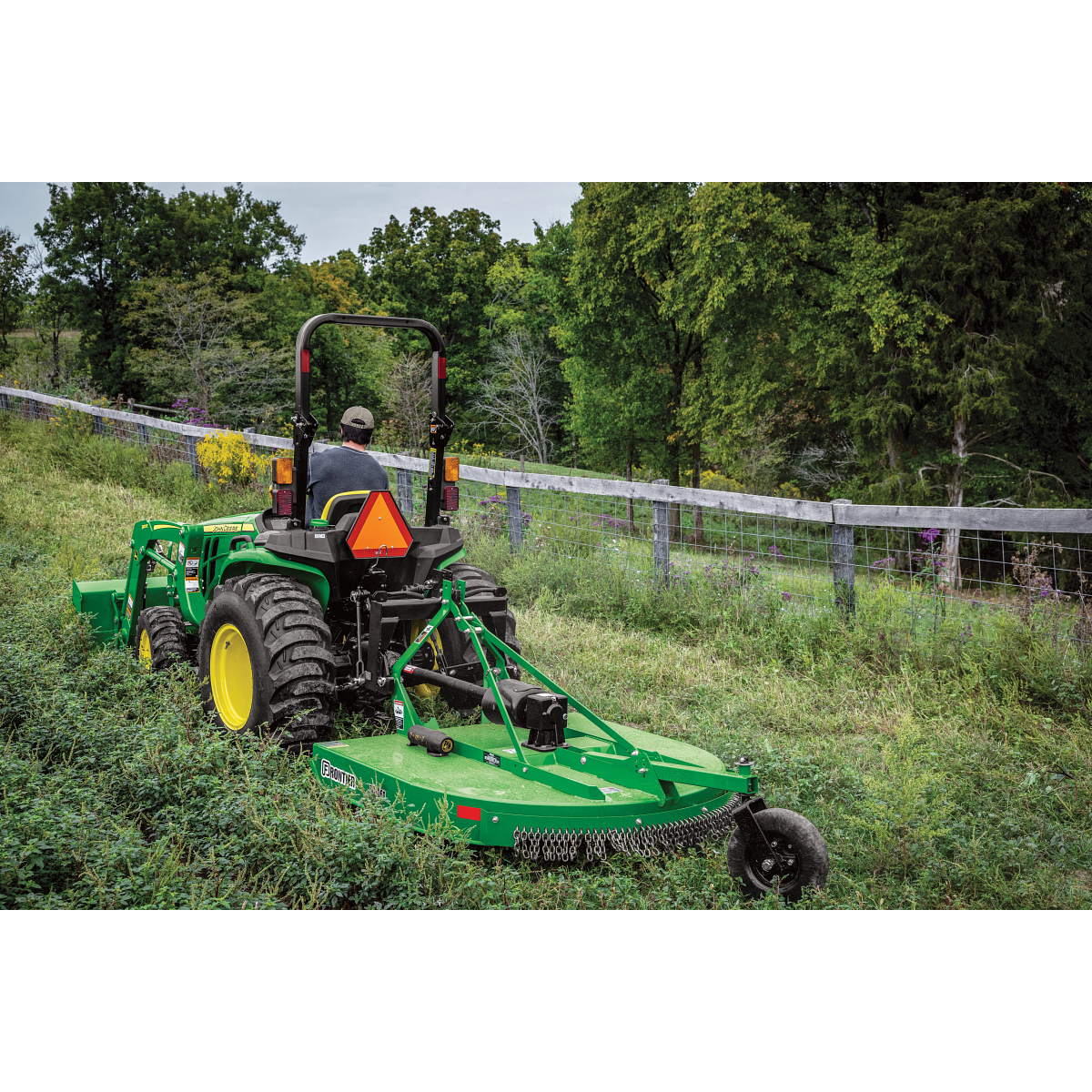 An operator on a green tractor with a rear-mounted mower cuts grass in a sloped field, with a wire fence and dense trees lining the background in a rural setting
