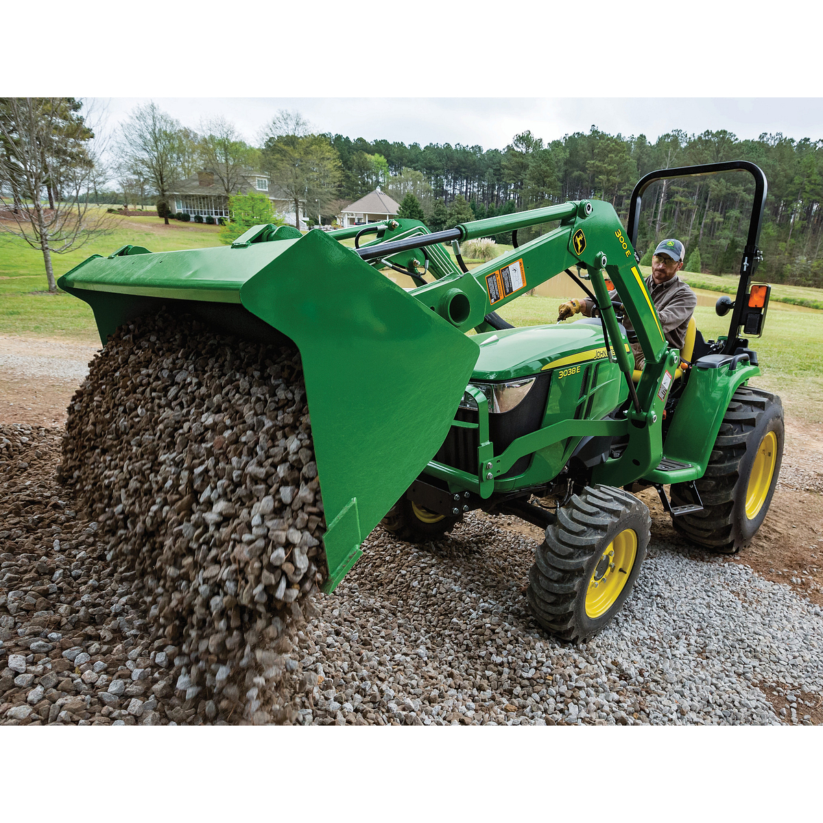 Green tractor with a front loader tipped forward, pouring a large load of gravel onto the ground and set in an open yard with grass, trees, and buildings visible in the background