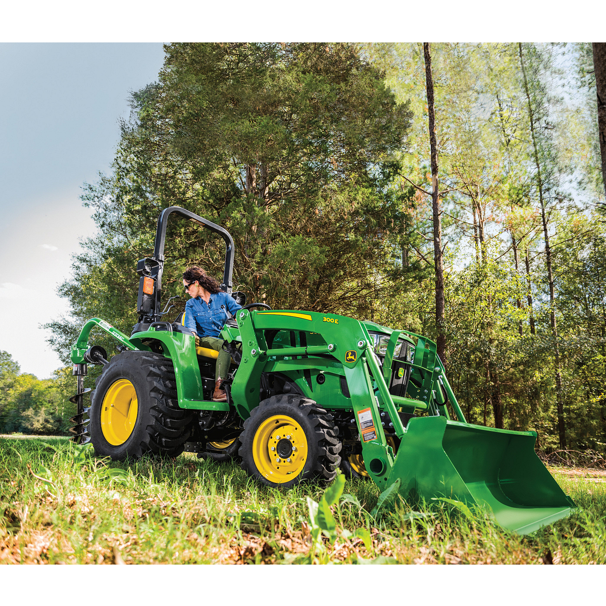 Person on a bright green tractor with a front loader attachment using a rear-mounted auger in a grassy field, with tall trees in the background under partly sunny conditions