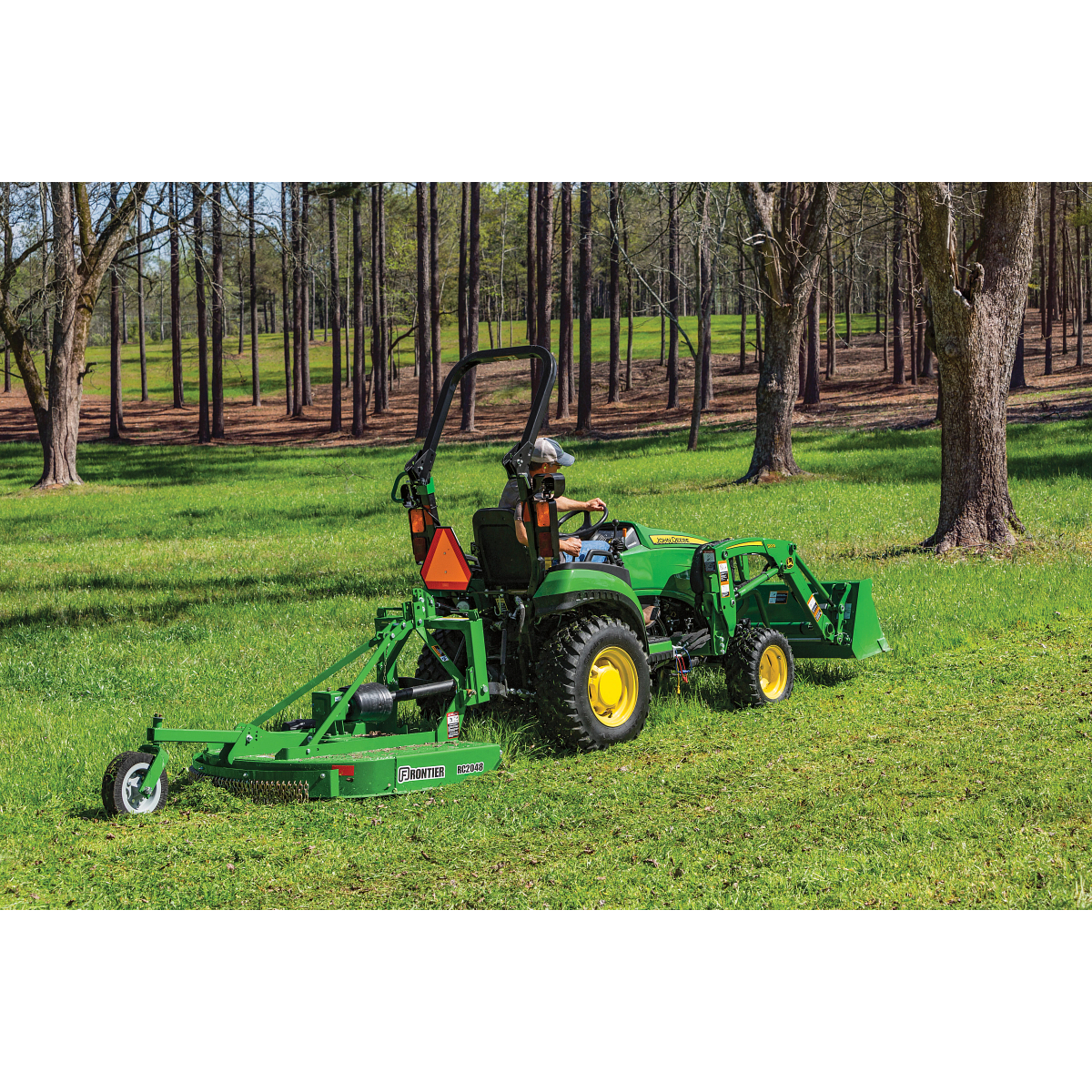 Green tractor with a front loader and rear mower attachment driving in a grassy field among evenly spaced trees in a wooded area under clear daylight