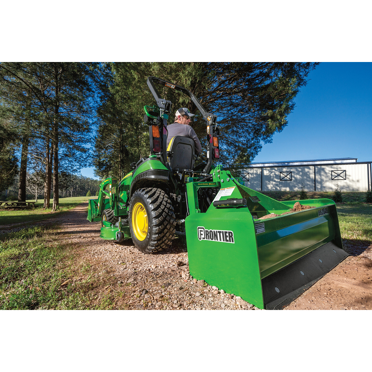 Green tractor equipped with a rear-mounted box blade leveling gravel on a path near trees and a light-colored building, with a person seated in the driver’s position