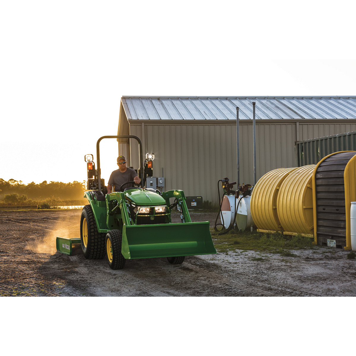 An operator on a green tractor with a front loader and rear land plane drives on a dirt path beside a metal building, with large tanks and equipment nearby at sunset