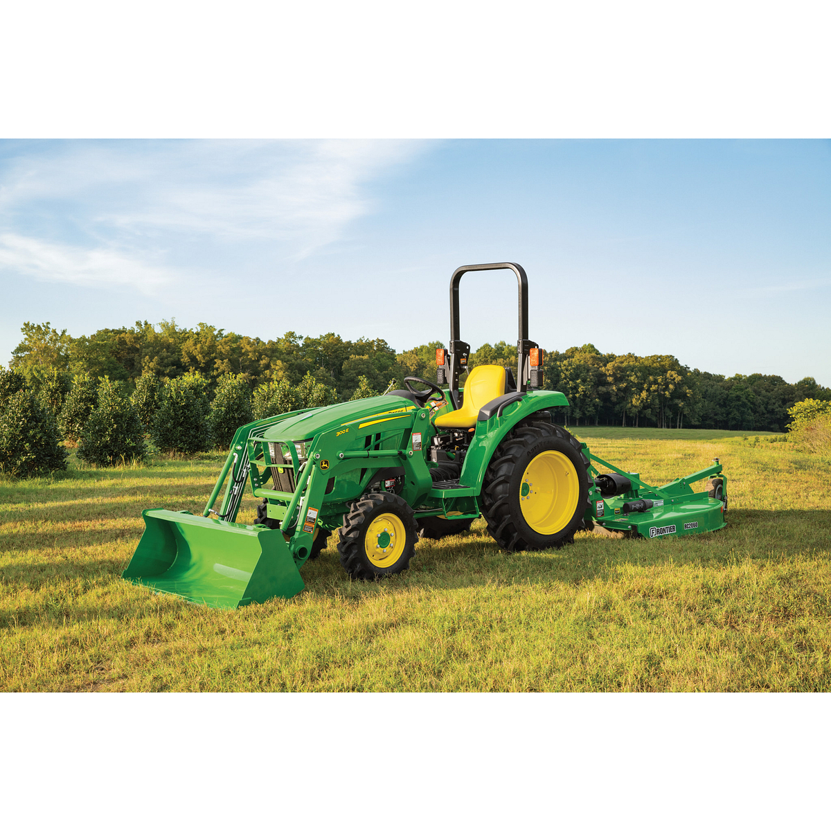 A green tractor with a front loader and rear rotary cutter sits in a grassy field under a clear sky, with trees lining the background and sun shining down