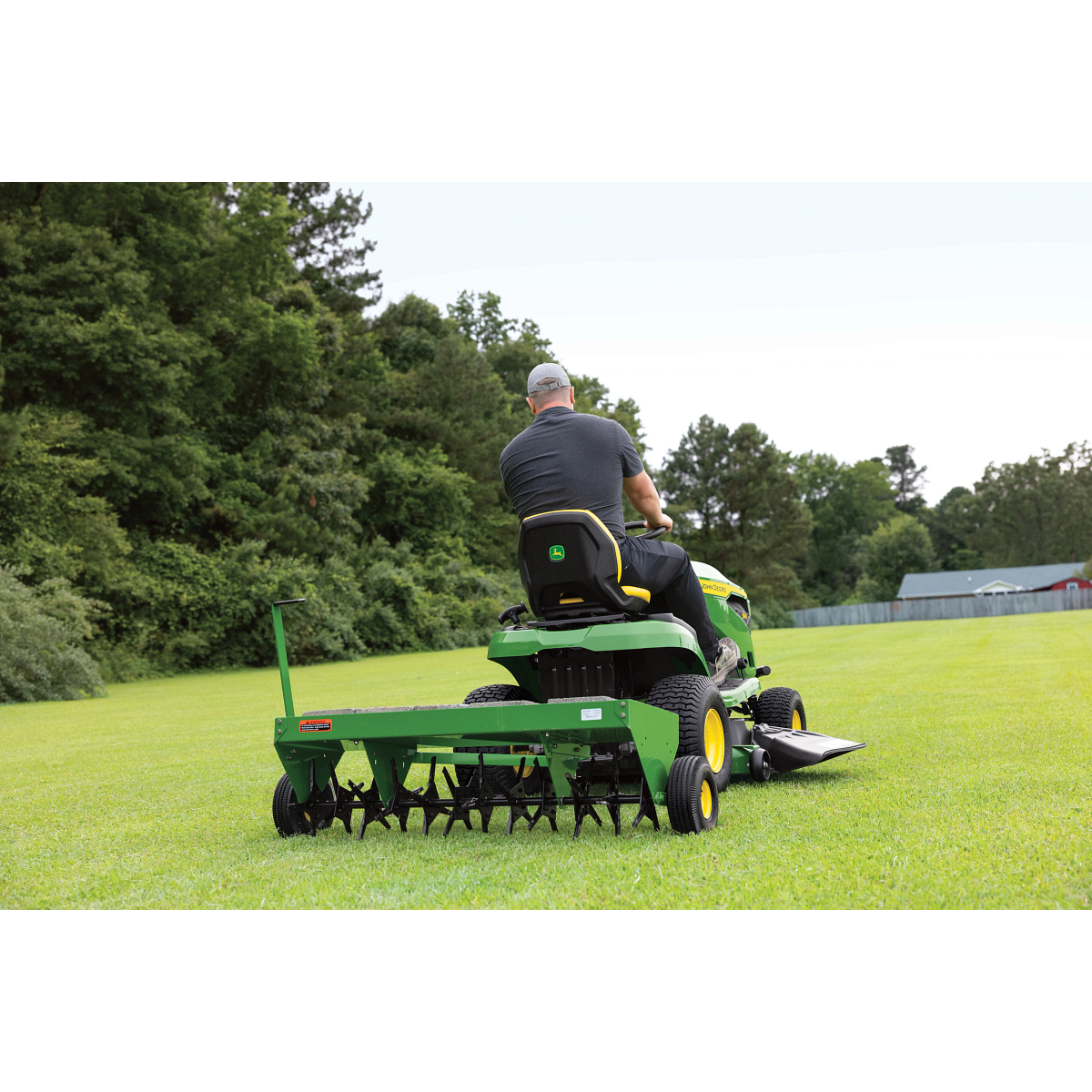 Rear view of an operator driving an S140 Lawn Tractor across a tree-lined lawn with attached aerator