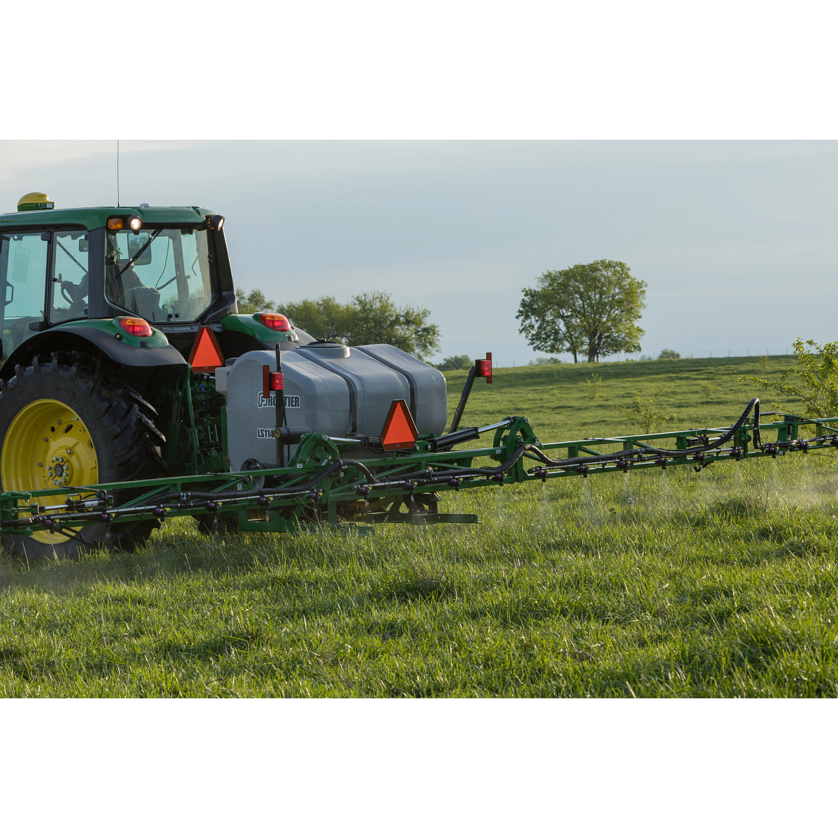 A tractor pulls a wide sprayer attachment across a grassy field, with a liquid tank mounted on the back and trees in the background under a clear sky