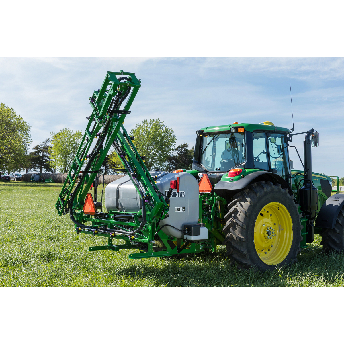 Agricultural tractor with yellow wheels parked on a grassy field, towing a folded boom sprayer with metal arms raised vertically and a large tank mounted behind the tractor under a clear blue sky.