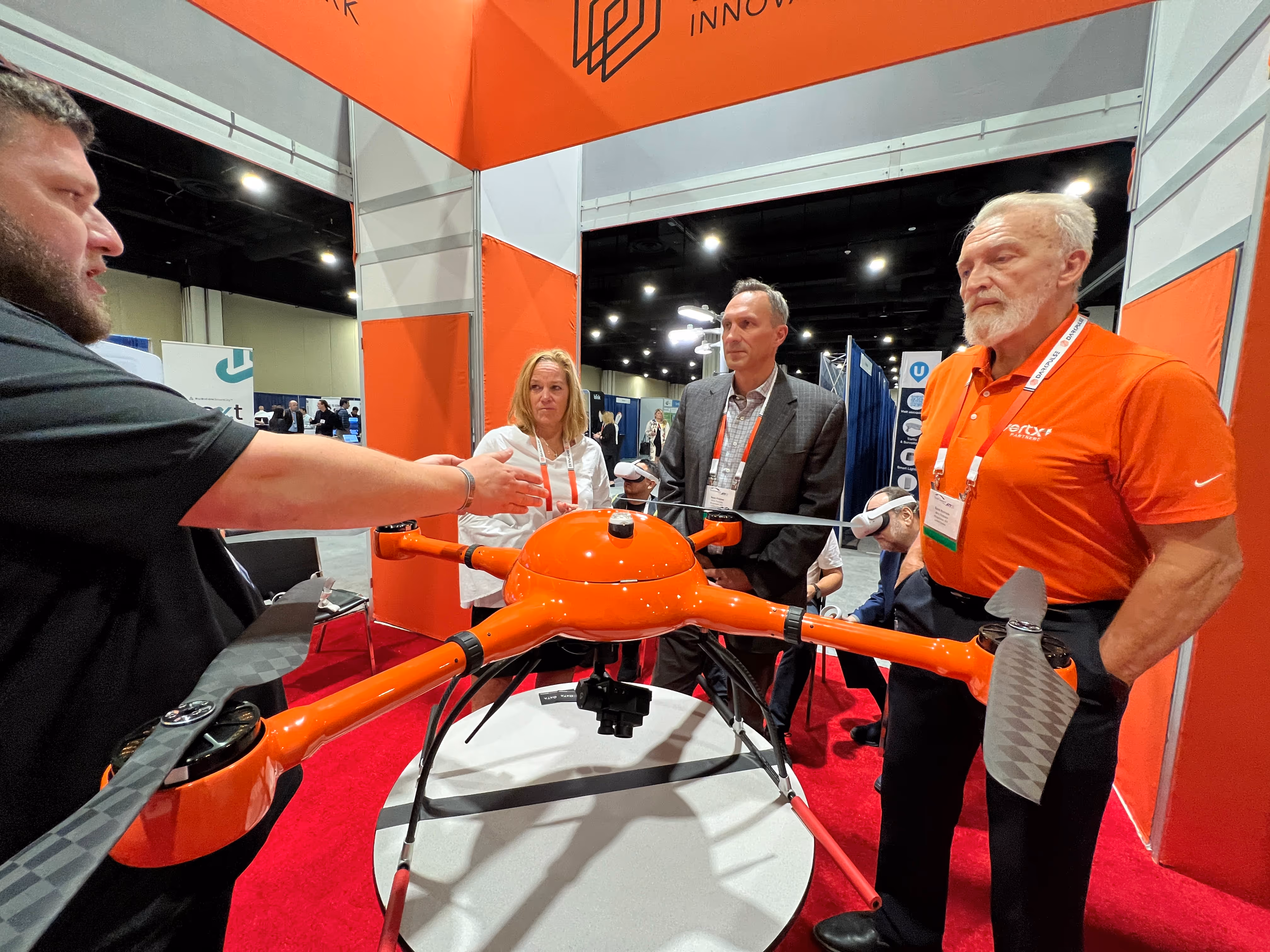 Four people gathered around a large orange drone displayed on a table at a convention or trade show.