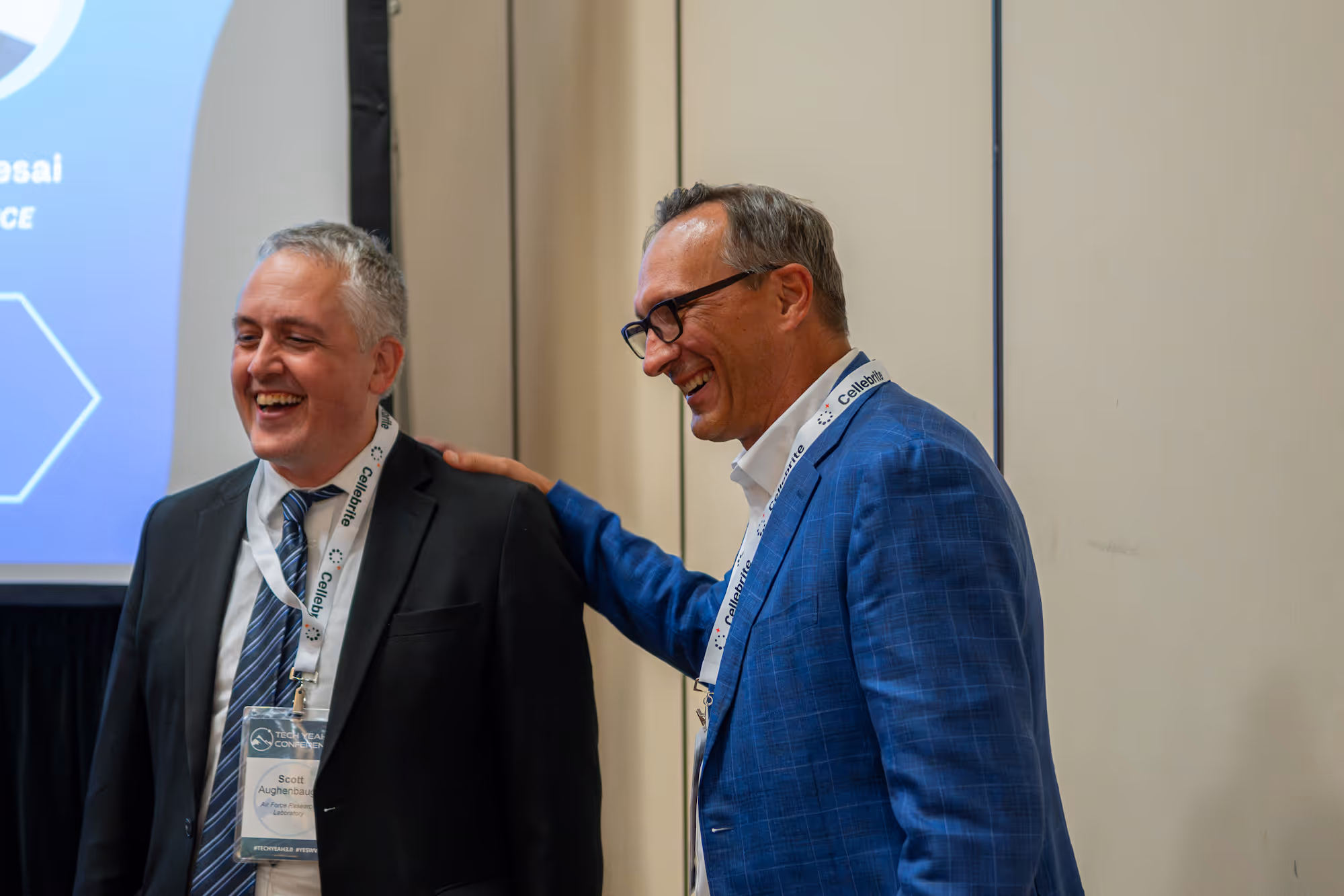 Two men smiling and laughing, one with a blue checkered suit and glasses has his hand on the other's shoulder.