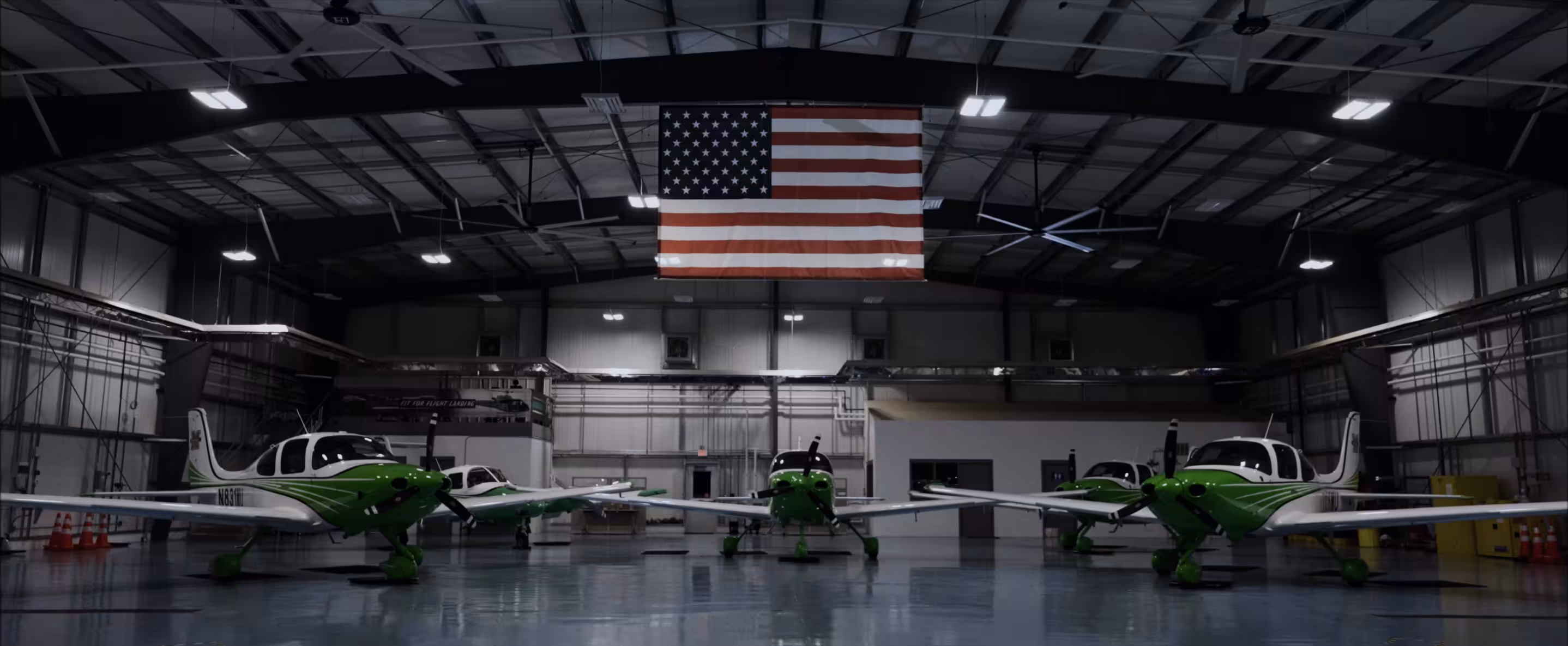 Four small green and white airplanes parked inside a large hangar with an American flag hanging from the ceiling.