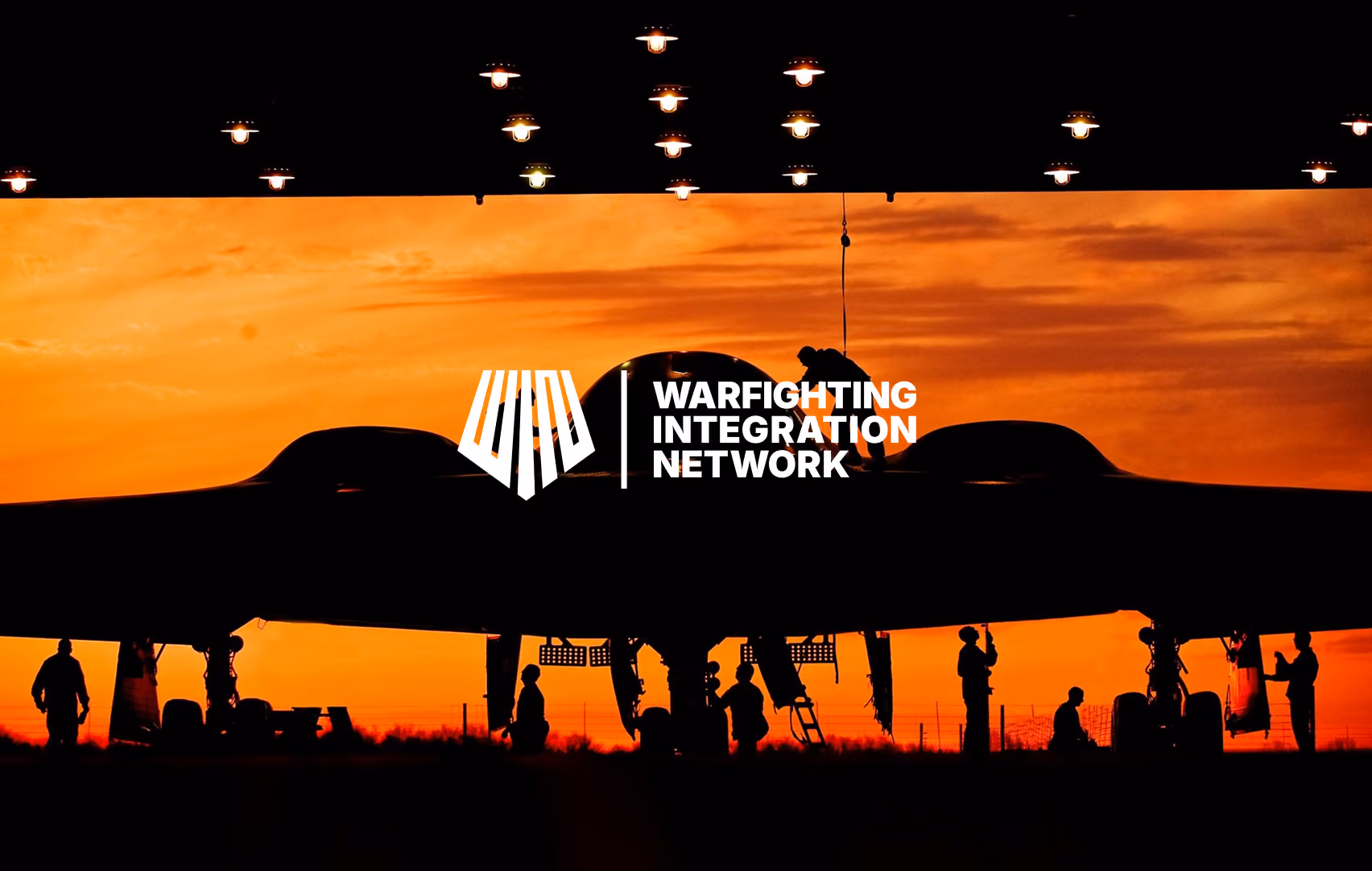Silhouettes of people servicing a large aircraft at sunset under hanging lights.