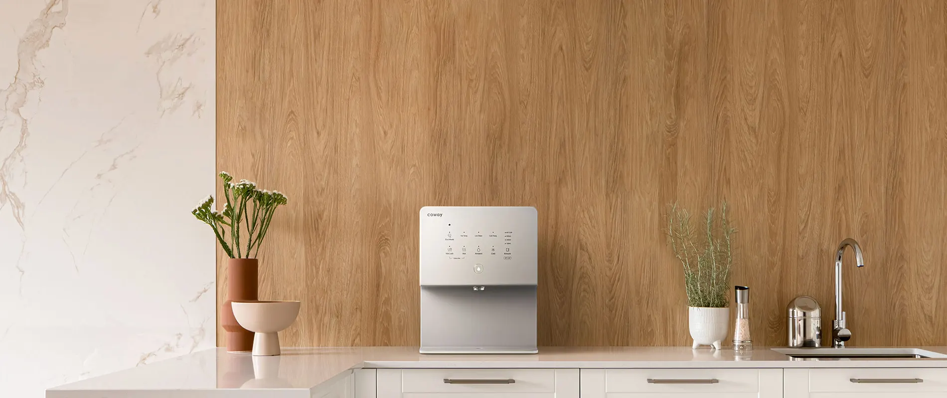 Modern kitchen countertop with Coway's CHP-6330L water purifier, potted plants, a salt grinder, and a chrome faucet against a wood-paneled wall.