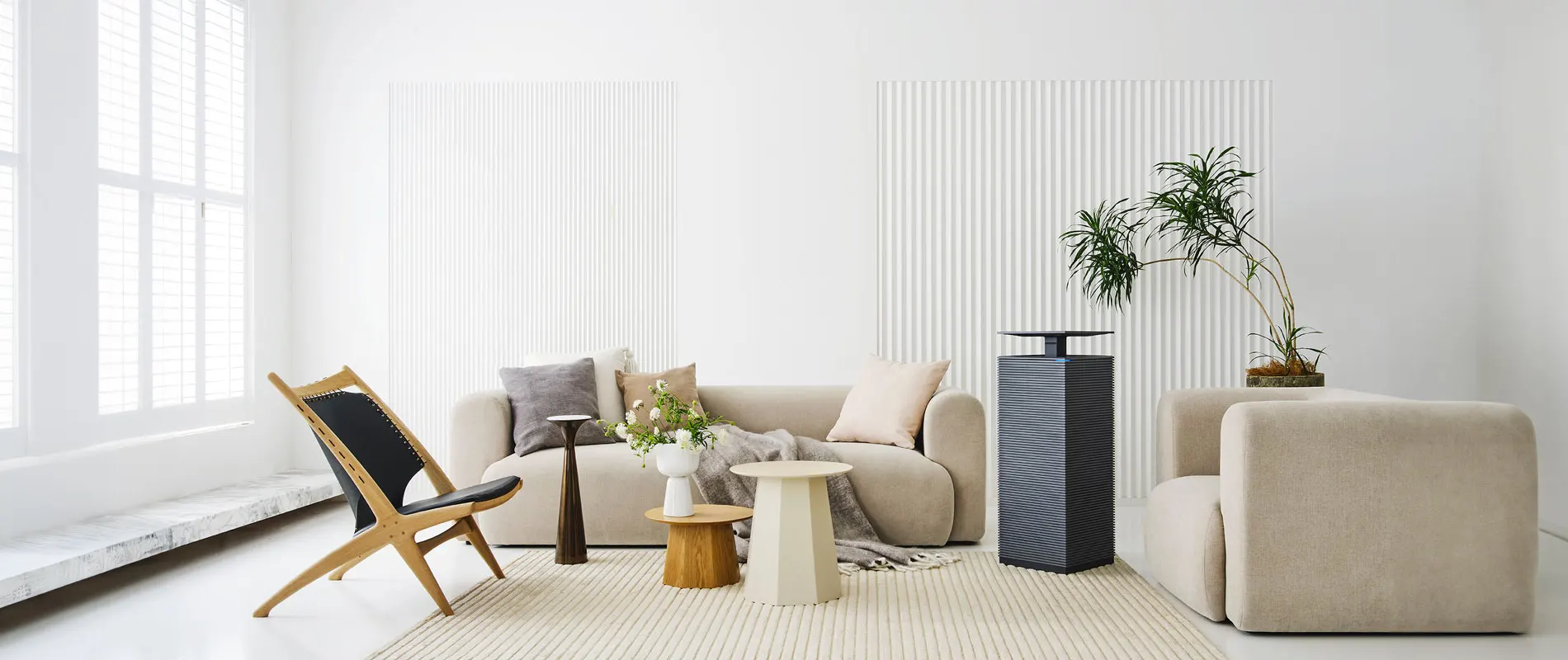 Minimalist living room with Coway's AP-2021A air purifier, beige sofas, black and wooden lounge chair, three small coffee tables, a floor plant, and large windows with white blinds.