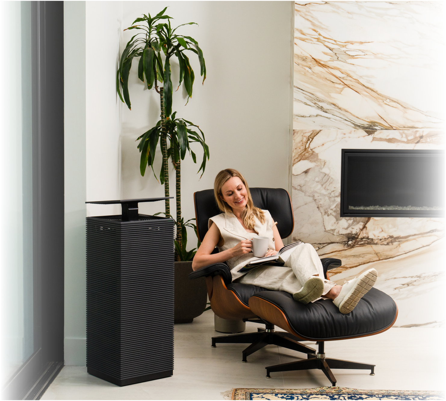 Woman sitting in a black leather lounge chair with wooden frame, next to Coway's AP-2021A air purifier, holding a mug and reading a book in a modern living room with a tall plant and marble fireplace.