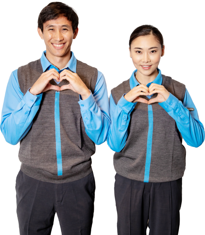 Smiling man and woman wearing blue shirts and gray vests forming heart shapes with their hands.
