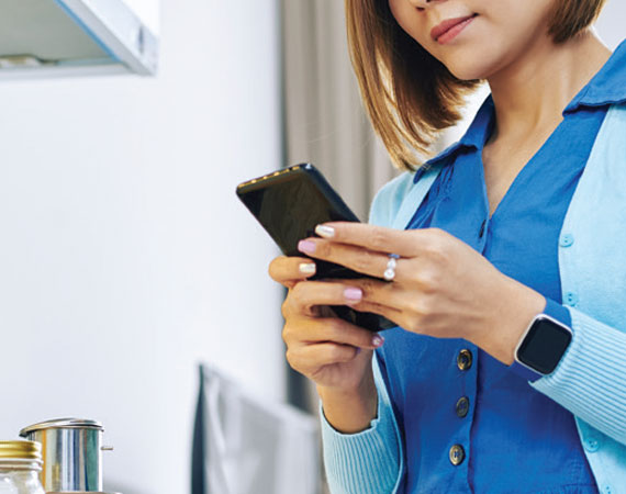 Woman wearing a blue cardigan and smartwatch, holding and looking at a smartphone indoors.