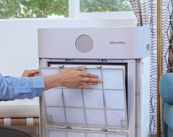 Person removing or cleaning the filter of a Coway air purifier in a living room setting.