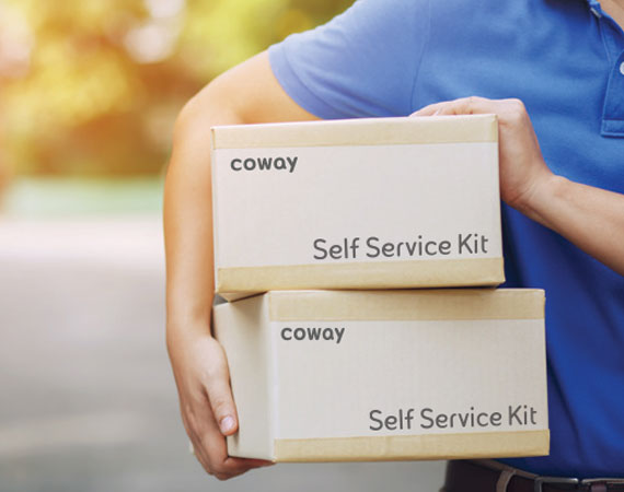 Person wearing a blue shirt holding two cardboard boxes labeled 'coway Self Service Kit' outdoors.