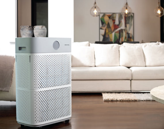 Modern white air purifier standing on wooden floor in front of a beige sofa in a well-lit living room.