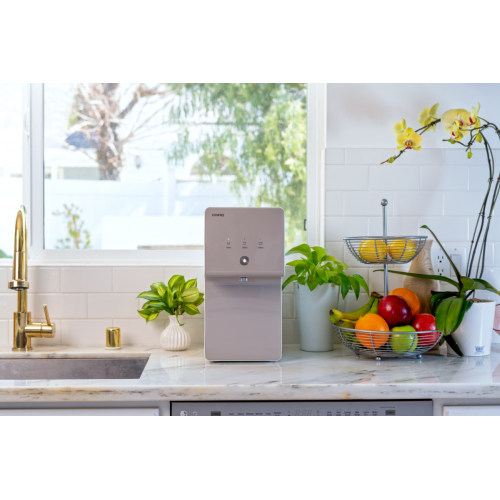Modern Coway P-6320L water purifier on a kitchen countertop next to a gold faucet, green plants, and a fruit basket with apples, bananas, and oranges.