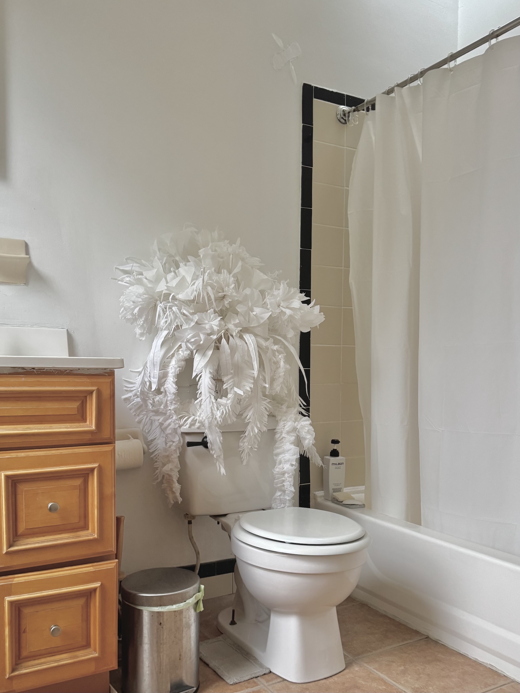 Bathroom with white toilet topped by a large white decorative flower arrangement, wooden cabinet on the left, metal trash can, and a bathtub with white shower curtain.