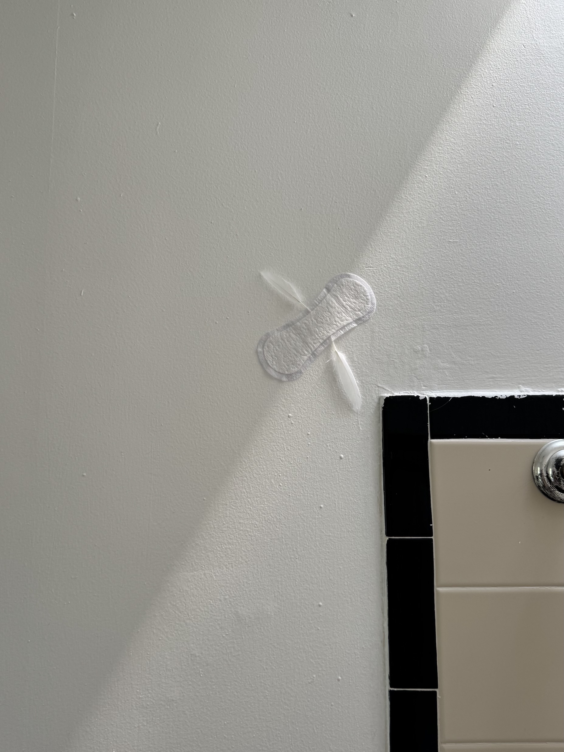 Small white sanitary pad on a white wall above black and beige tiles.