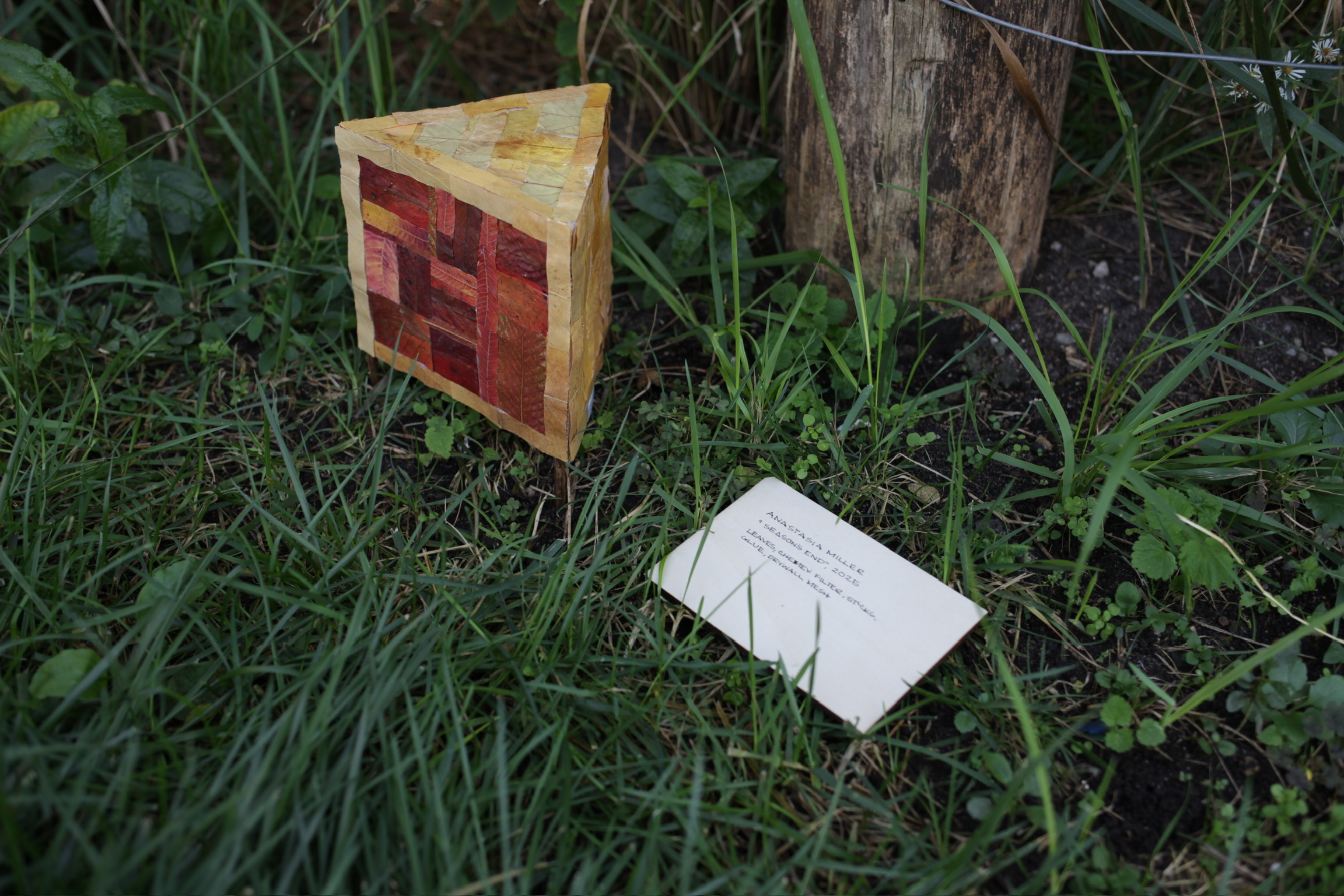 Small triangular lantern with red and yellow leaf patterns planted in grassy soil next to a wooden stump with an art label nearby.