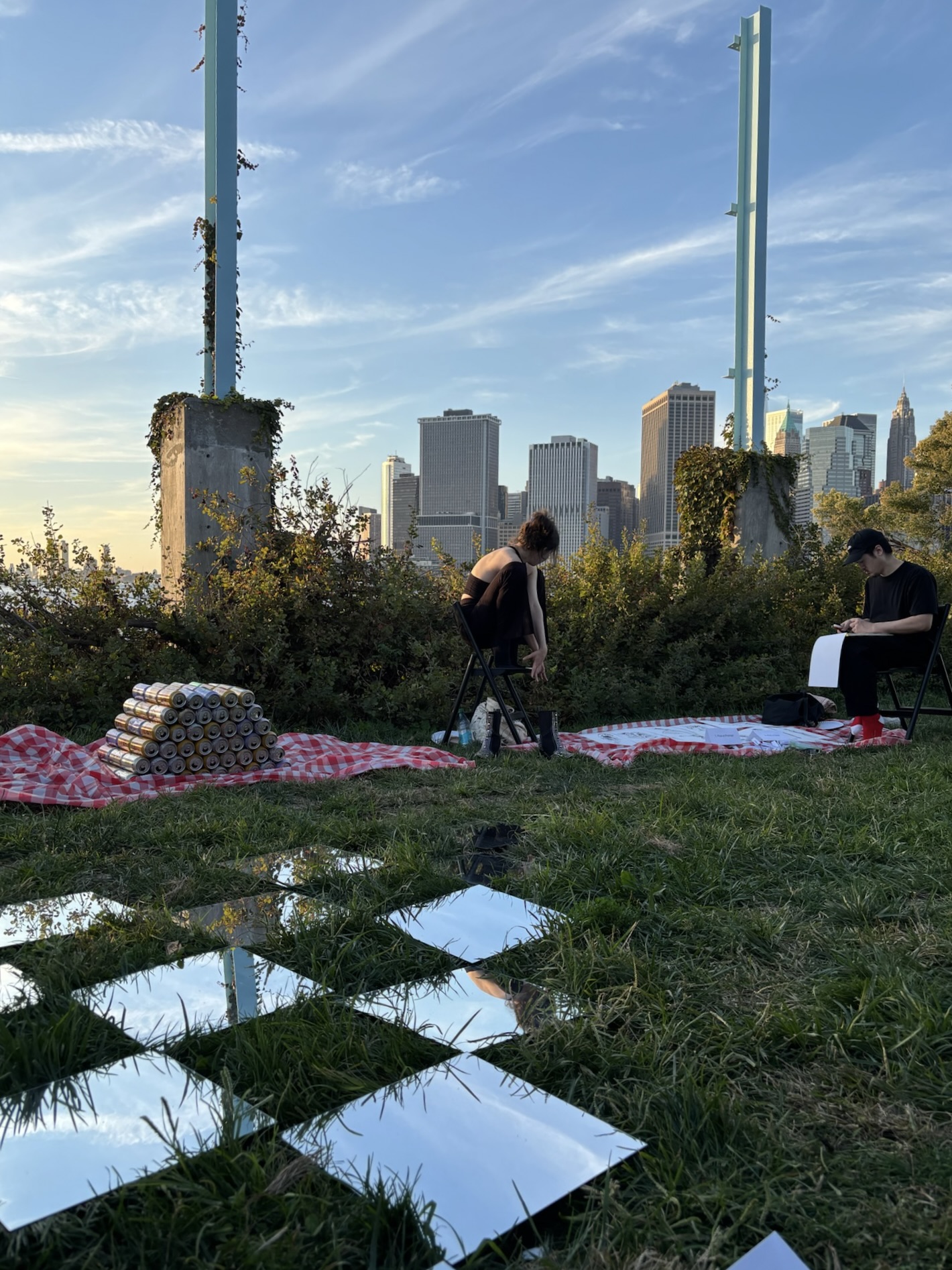 Two people seated outdoors on folding chairs with red checkered blankets, a mastaba of cans, and square mirrors on the grass, with a city skyline in the background at sunset.