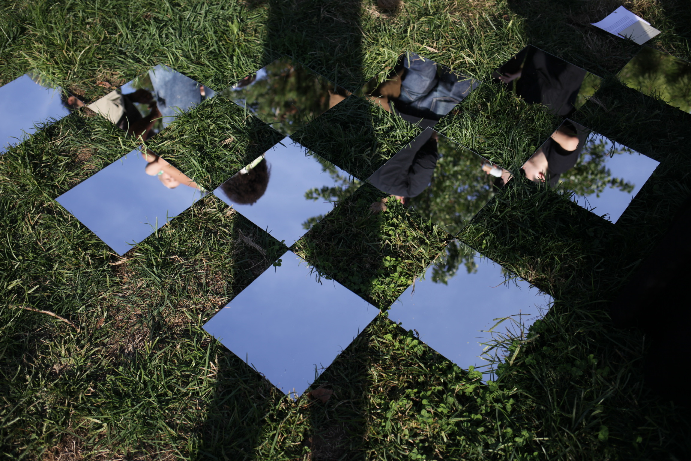 Square mirrors laid on grass reflecting parts of people, blue sky, and tree branches.