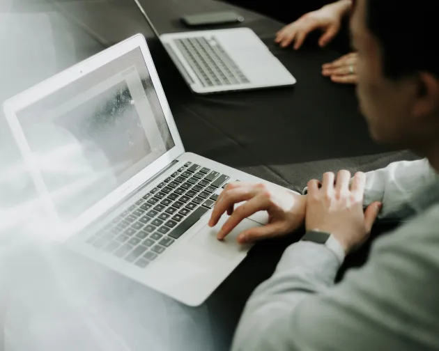 A person works on a laptop at a table, with another laptop visible in the background, focusing on the screen.