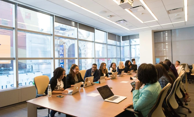 A diverse group of professionals engaged in a meeting around a large conference table with laptops, in a bright, modern office space.
