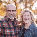 A couple stands close together outdoors, surrounded by golden autumn foliage, smiling and dressed in casual outfits.