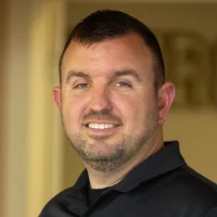 A person in a black polo shirt stands in a warmly lit interior, slightly turned to the side, with a neutral expression.