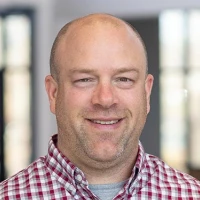 A person in a red and white checked shirt stands indoors, with a blurred face and a neutral expression. Soft lighting in the background.