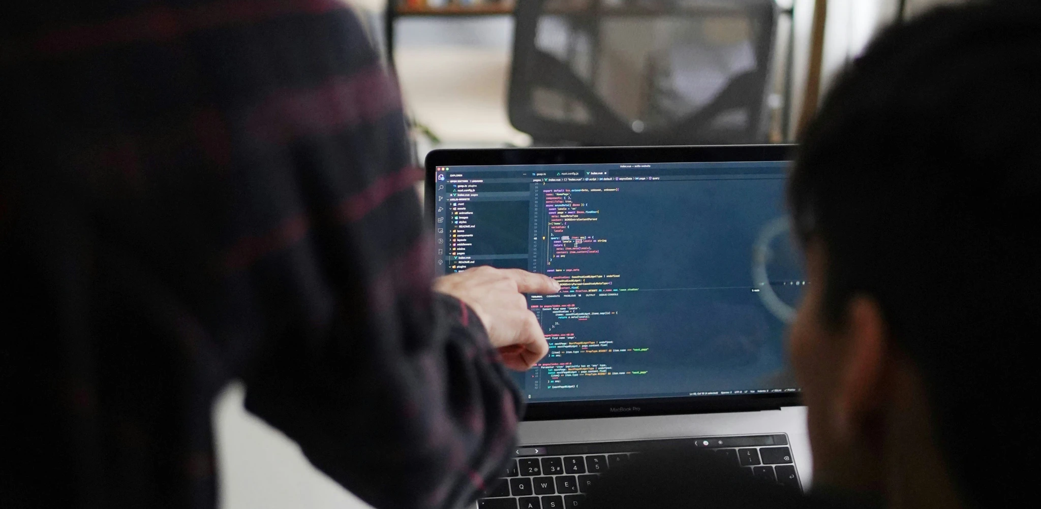 A person points at code on a laptop screen while another person observes, engaged in a programming discussion.