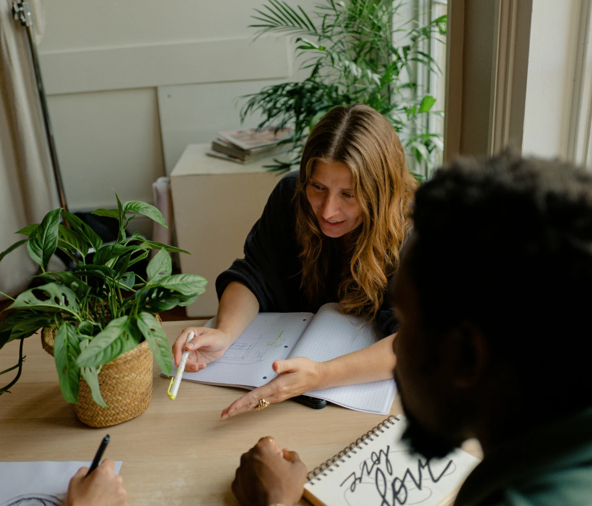 A group discusses ideas at a table, surrounded by plants and notebooks, with one person gesturing while others listen intently.