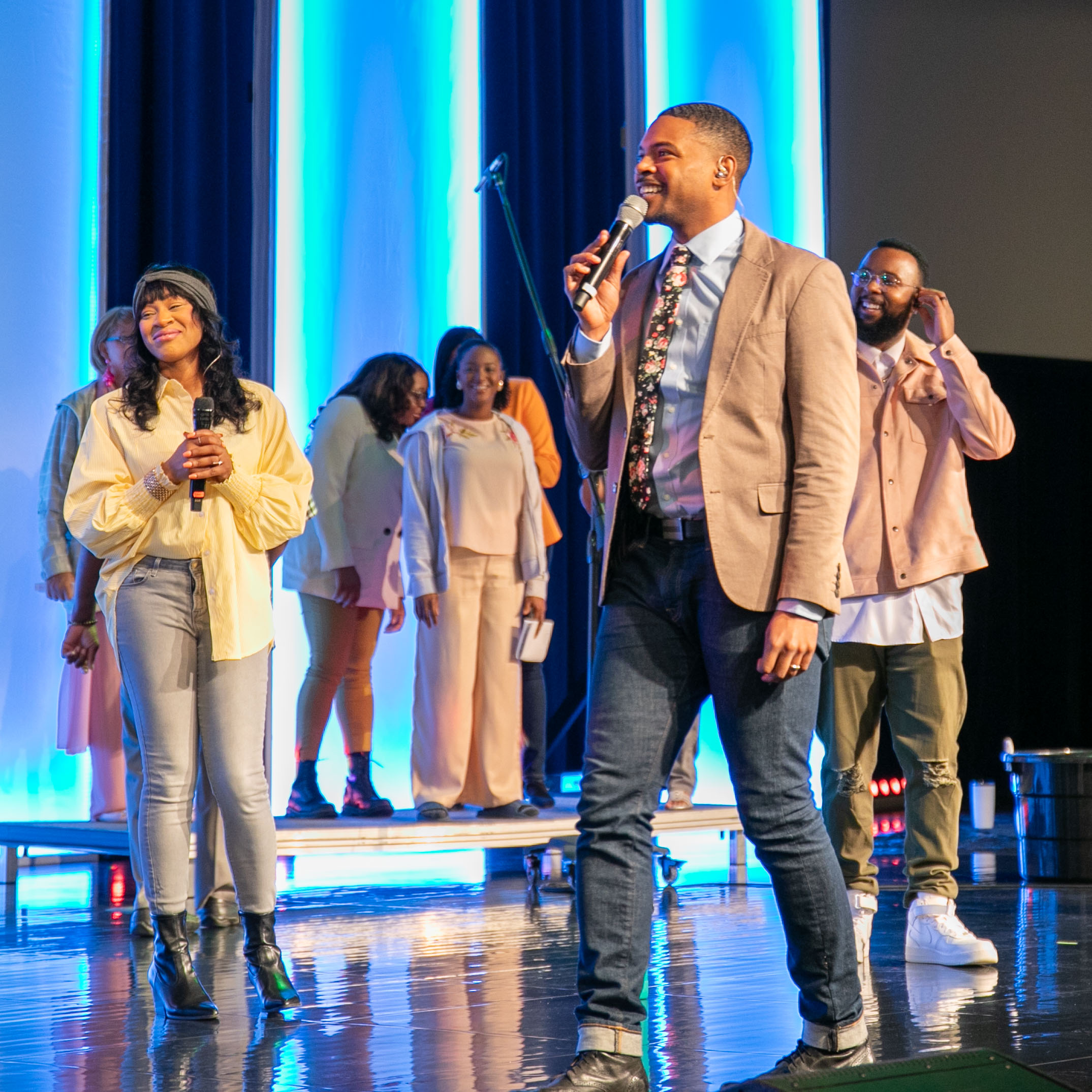 Two people singing on stage with a group of backup singers behind