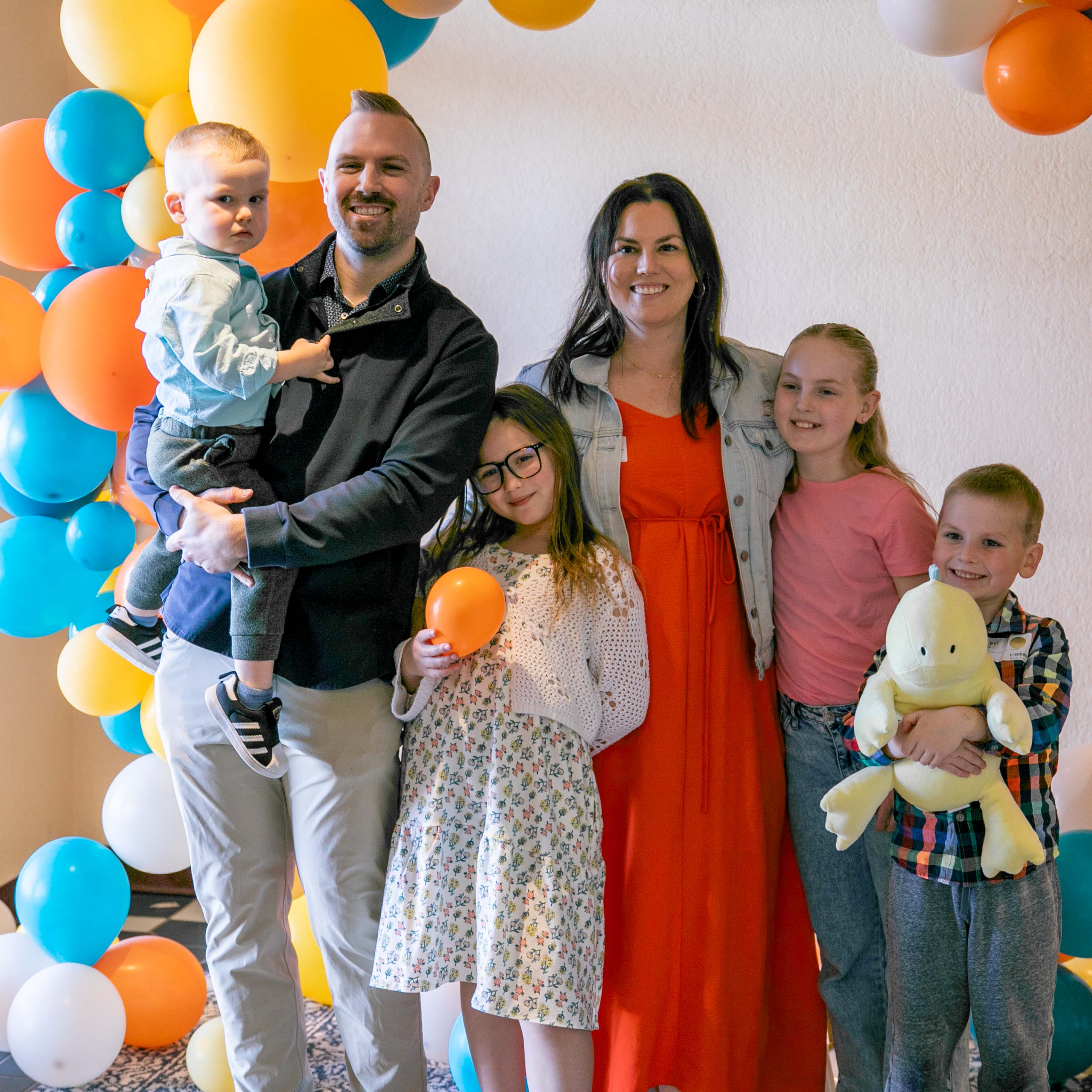 Smiling family of six including two adults and four children standing in front of a colorful balloon arch.