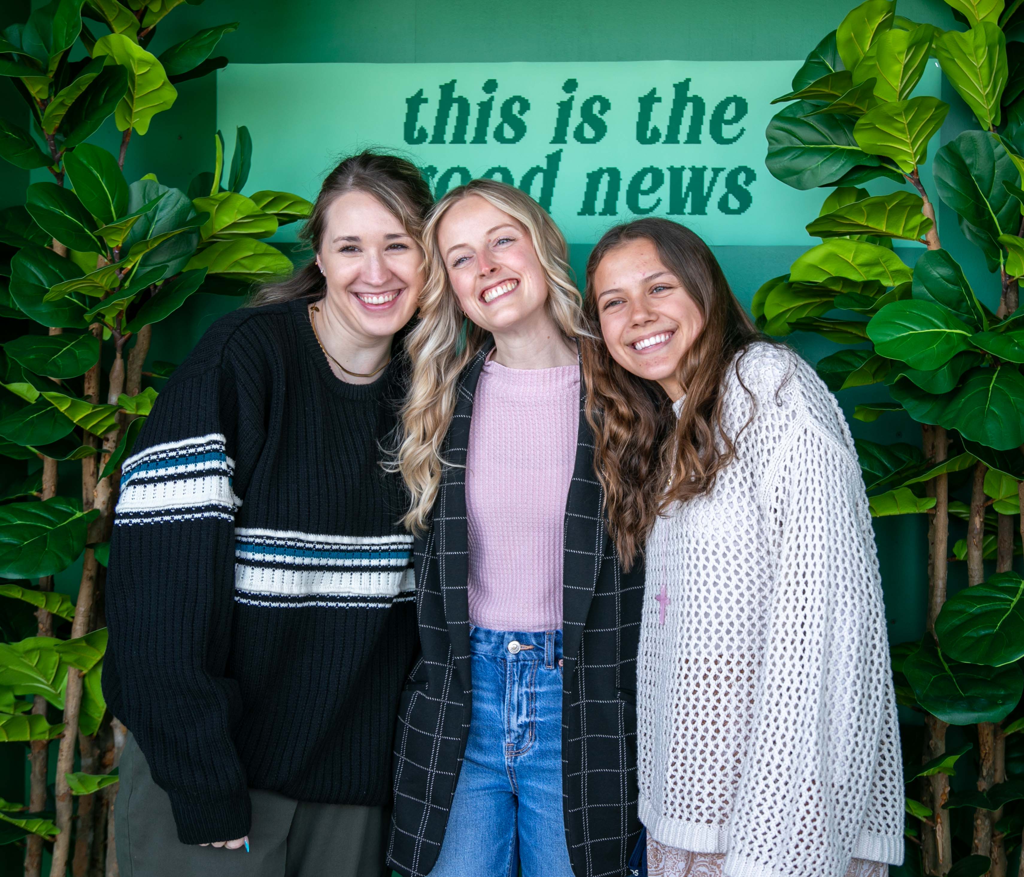 Three smiling young women standing closely together in front of a turquoise wall with the text 'this is the good news', surrounded by green potted plants.