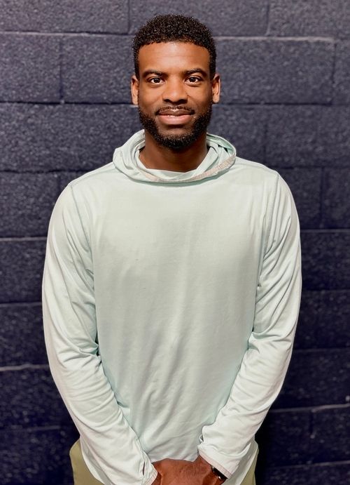 Smiling man with trimmed beard wearing a light hoodie standing against a dark brick wall.