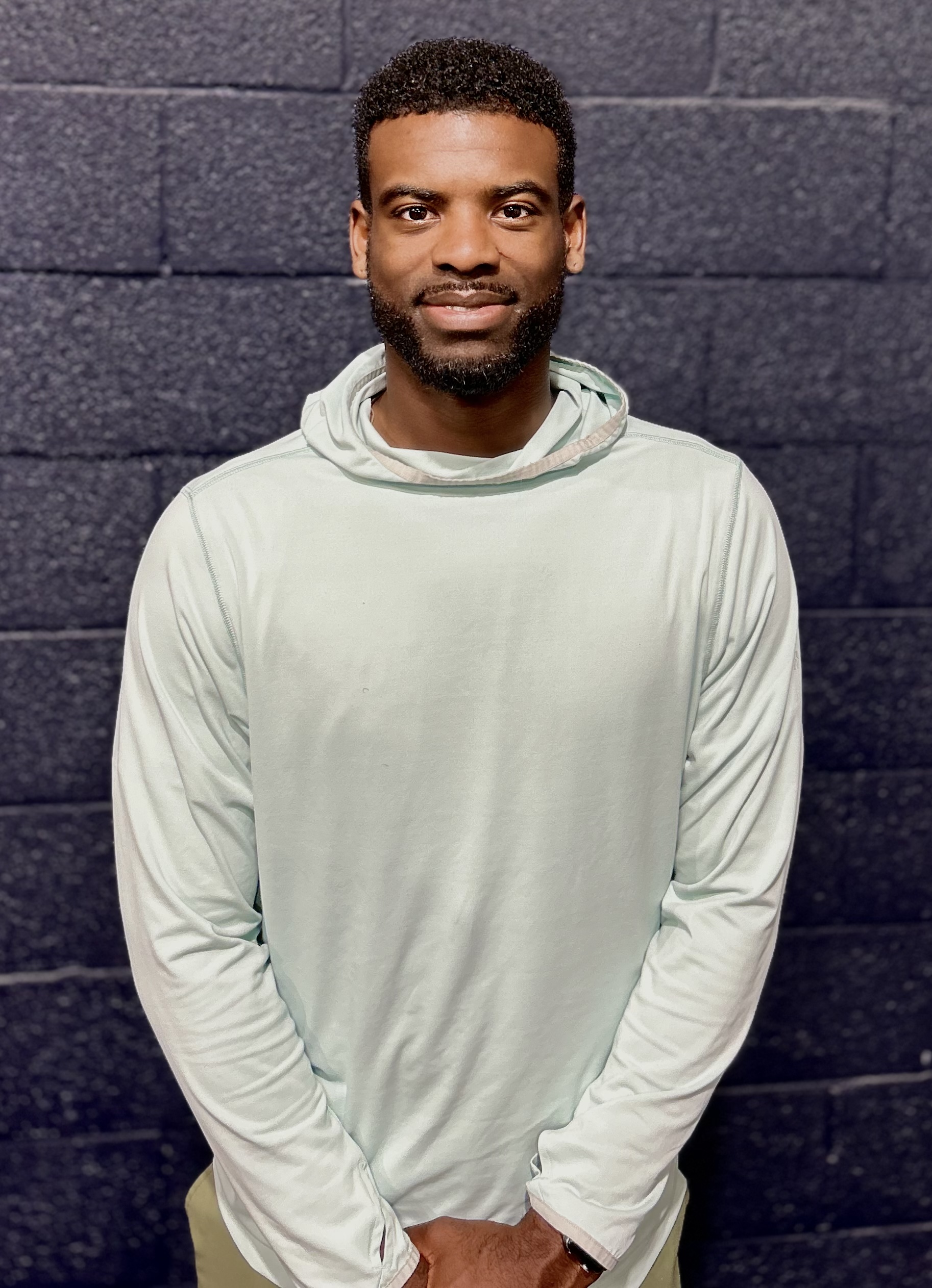Smiling man with trimmed beard wearing a light hoodie standing against a dark brick wall.