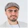 Man with a beard wearing a gray flat cap, light shirt, and beige jacket smiling against a white background.
