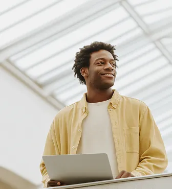 Smiling young man holding a laptop and looking to the side in a bright indoor space with a skylight ceiling.