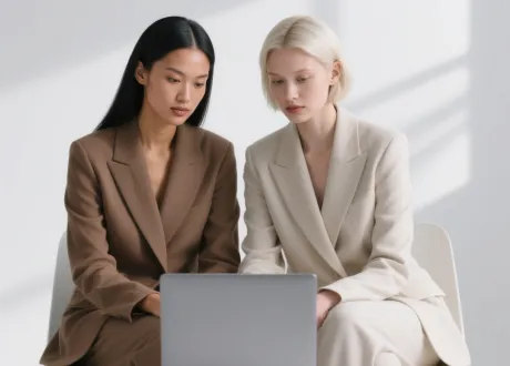 Two women in brown and beige suits sitting side by side, focused on a laptop in front of them.