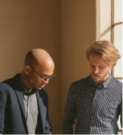 Two men standing closely, looking down and engaging in discussion near a sunlit window.