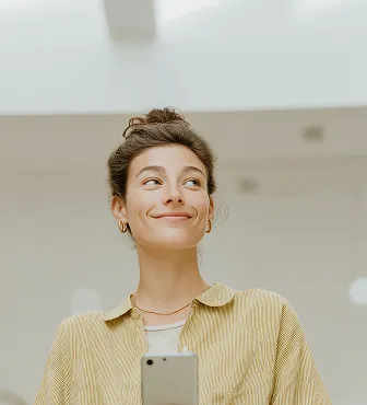 Young woman with hair in a bun, wearing a yellow striped shirt, looking to the side and holding a smartphone.