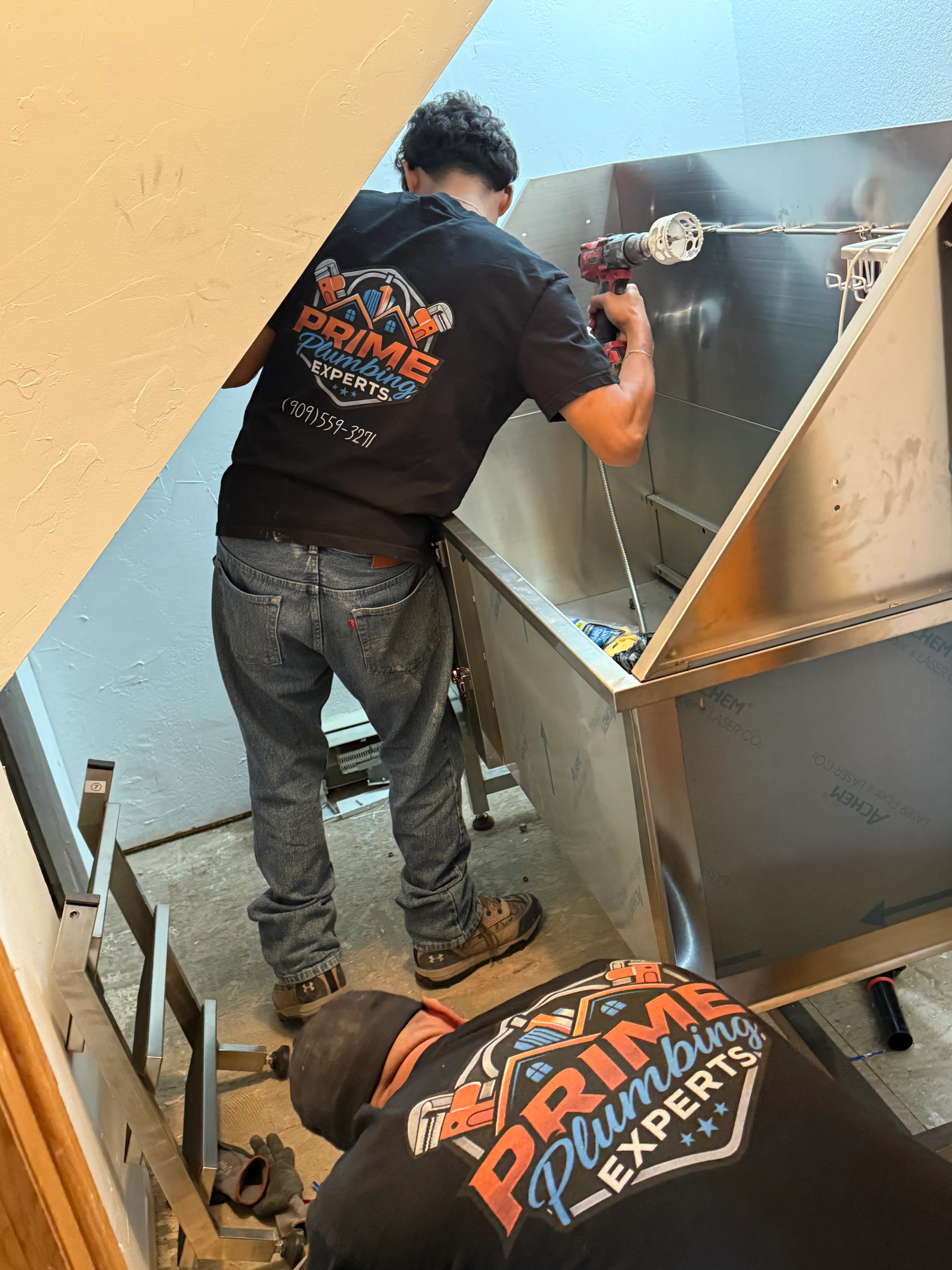 Two plumbers wearing black Prime Plumbing Experts t-shirts installing or working on a large stainless steel sink or fixture in a corner space.