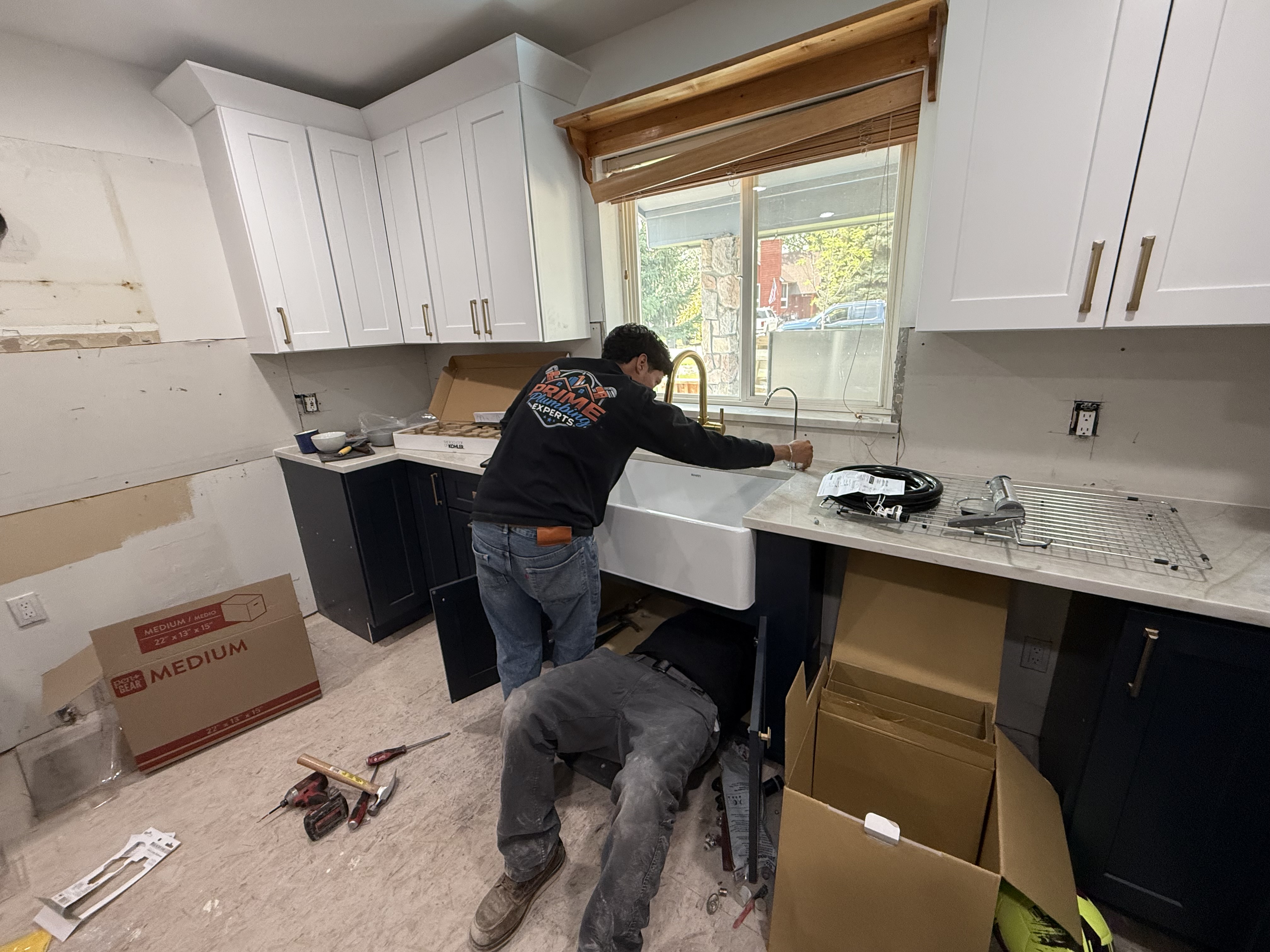 Two workers installing a white farmhouse kitchen sink under a window in a kitchen with white upper and dark lower cabinets.